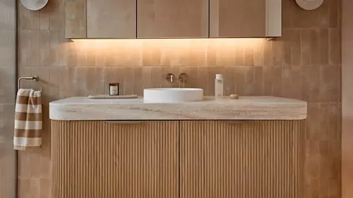 Modern bathroom vanity with white vessel sink, marble countertop, and fluted wood cabinet under beige tile wall.