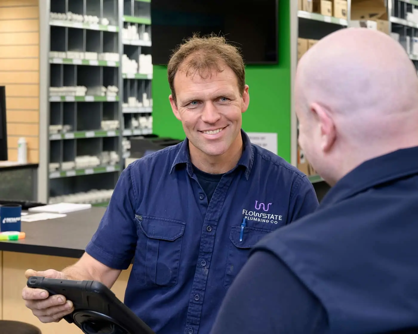 Two men in blue work uniforms talking in a plumbing supply store with shelves of products in the background.