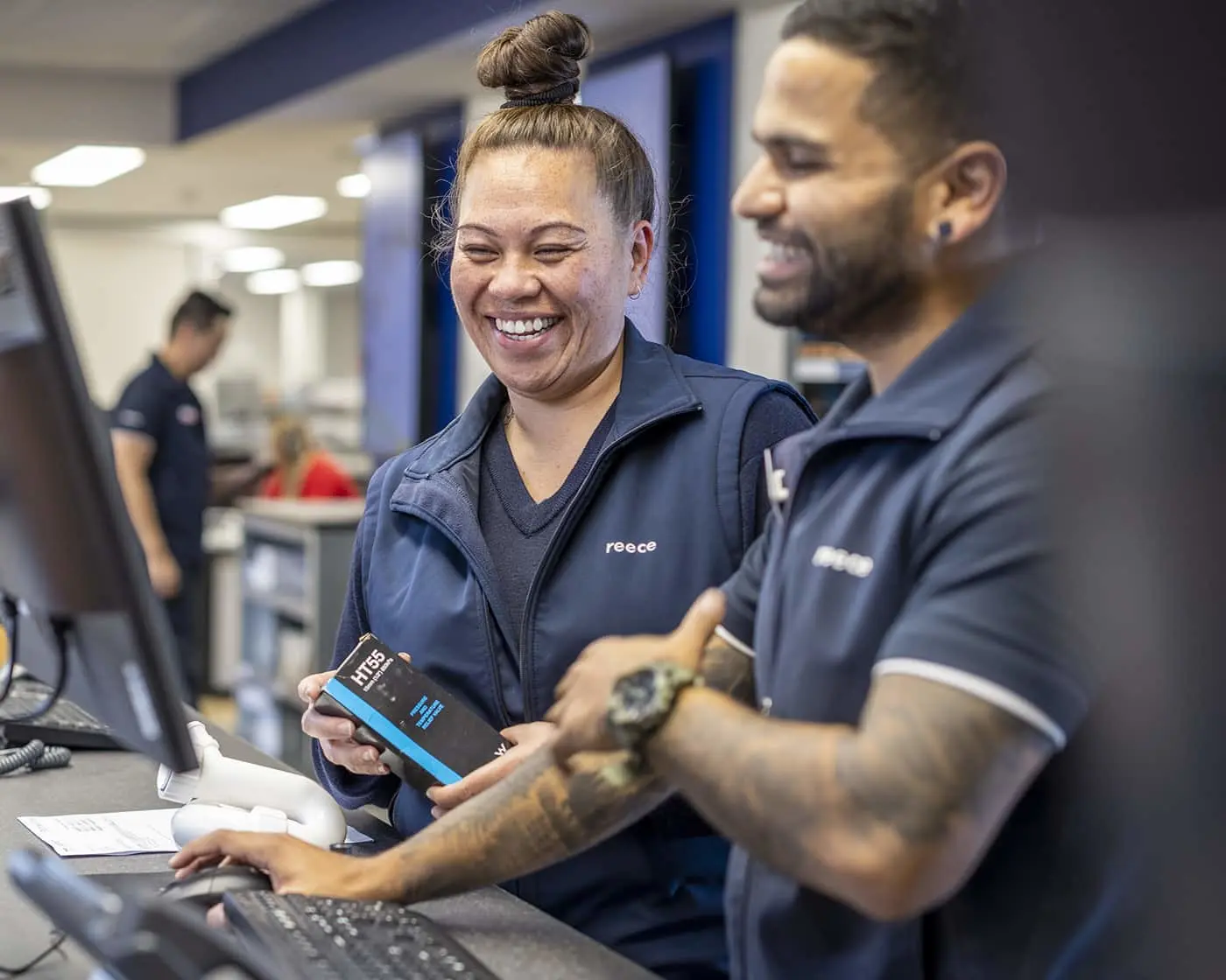 Two employees in navy uniforms smiling while working at a counter with a computer and phone.