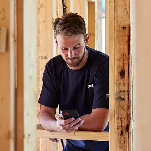 Man in dark blue shirt looking at phone inside wooden frame construction of unfinished building.