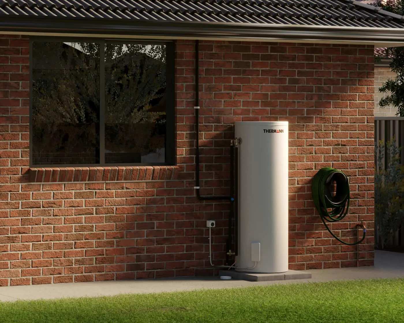 White cylindrical water heater installed outside a red brick house with a coiled green hose mounted on the wall.