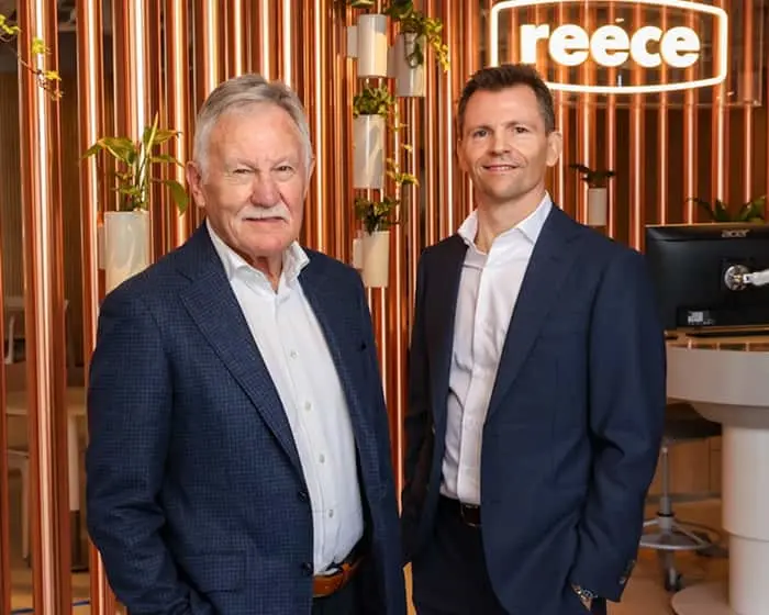 Two men in business suits standing in an office with copper-colored vertical panels and plants, with a reece logo visible.
