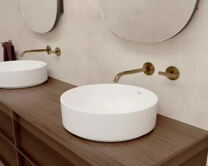 Modern bathroom with two white round vessel sinks on wood counter, brass wall-mounted faucets, and round mirrors.