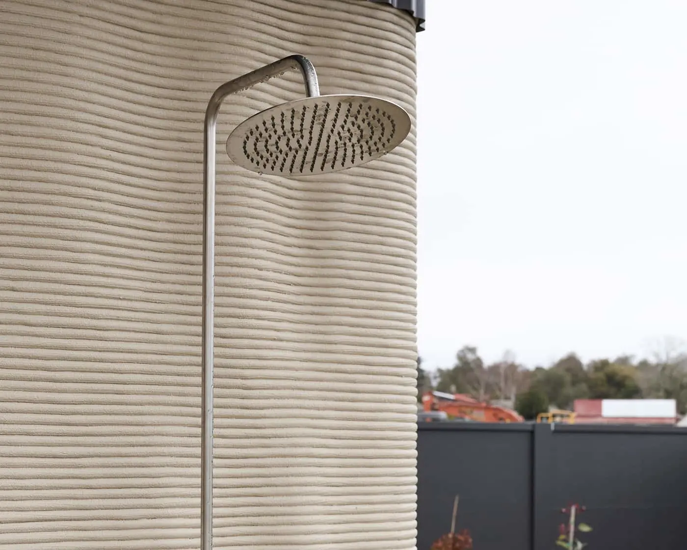 Outdoor shower head mounted on textured beige wall with blurred background of trees and buildings.