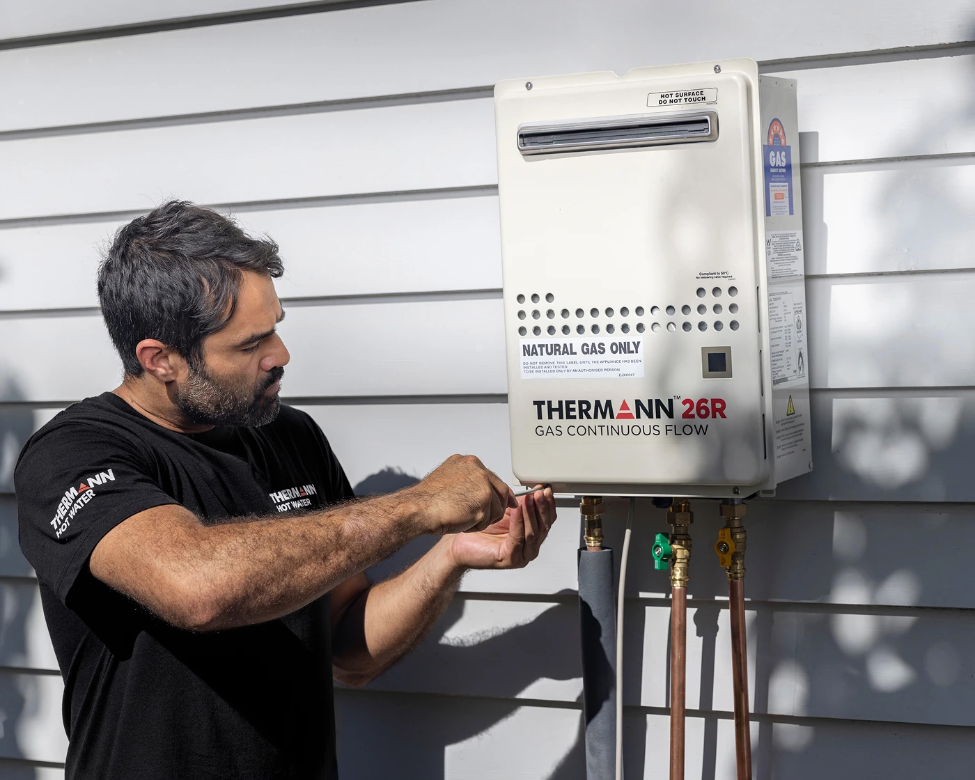 Technician installing a Thermann 26R natural gas continuous flow water heater on an exterior wall.