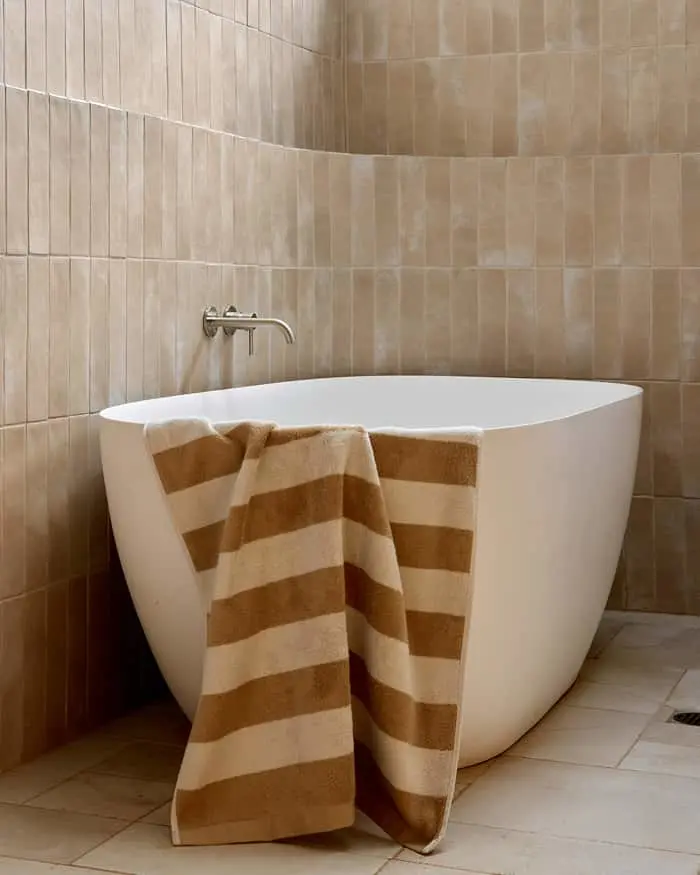 White freestanding bathtub with beige striped towel draped over the side against tan tiled walls