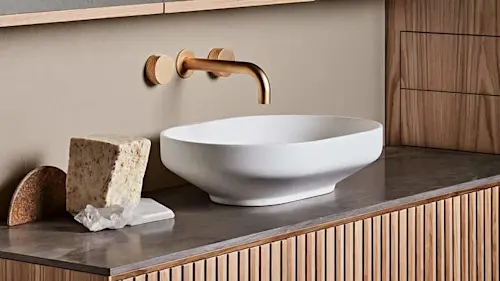 White vessel sink on gray countertop with brass wall-mounted faucet and wood cabinetry