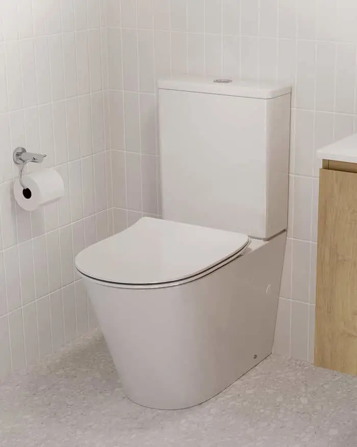 White toilet with closed lid in corner of bathroom with white tiled walls and toilet paper holder