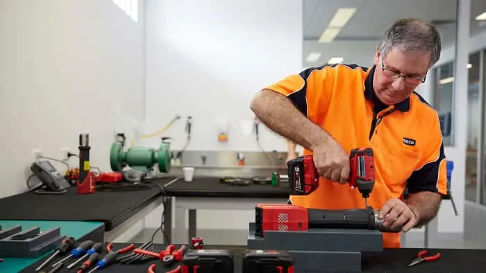 Man in orange safety shirt using a red power drill at a workbench with tools in a workshop setting.