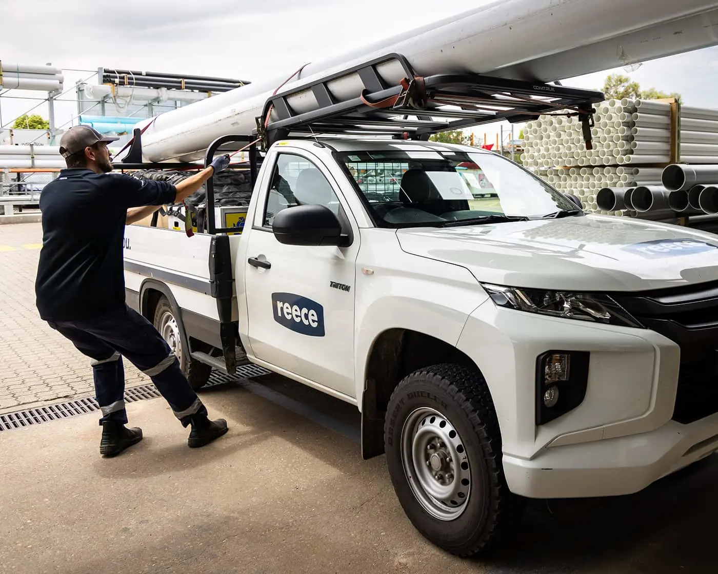 Worker loading PVC pipes onto roof rack of white Reece utility truck at supply yard.
