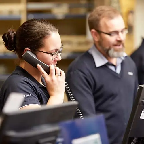 Woman with glasses talking on phone at desk with male colleague in background wearing dark polo shirts