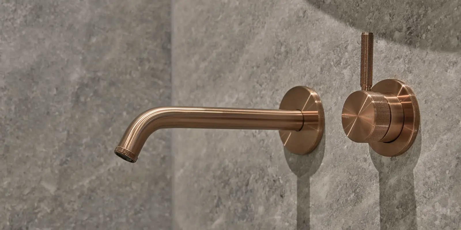 Bronze-colored wall-mounted faucet with textured knobs against a gray concrete wall.