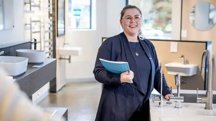 Woman in dark blazer holding blue folder smiling in bathroom showroom with sinks and fixtures displayed