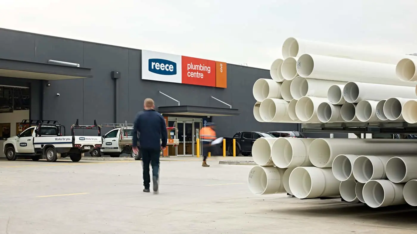 Person walking toward Reece plumbing centre with white service vehicles parked outside and stacked white pipes in foreground.