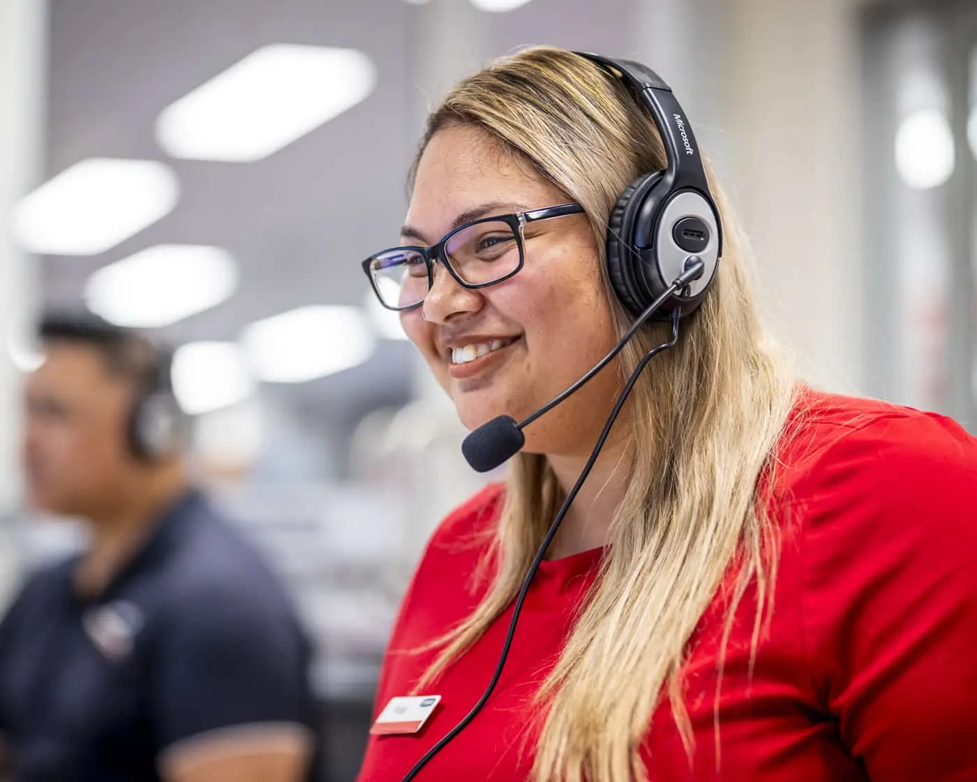 Woman wearing glasses and headset with microphone, smiling in red shirt in office setting