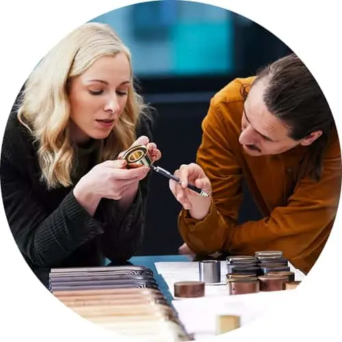 Two women examining a small circular sample, with material swatches displayed on a table in front of them.