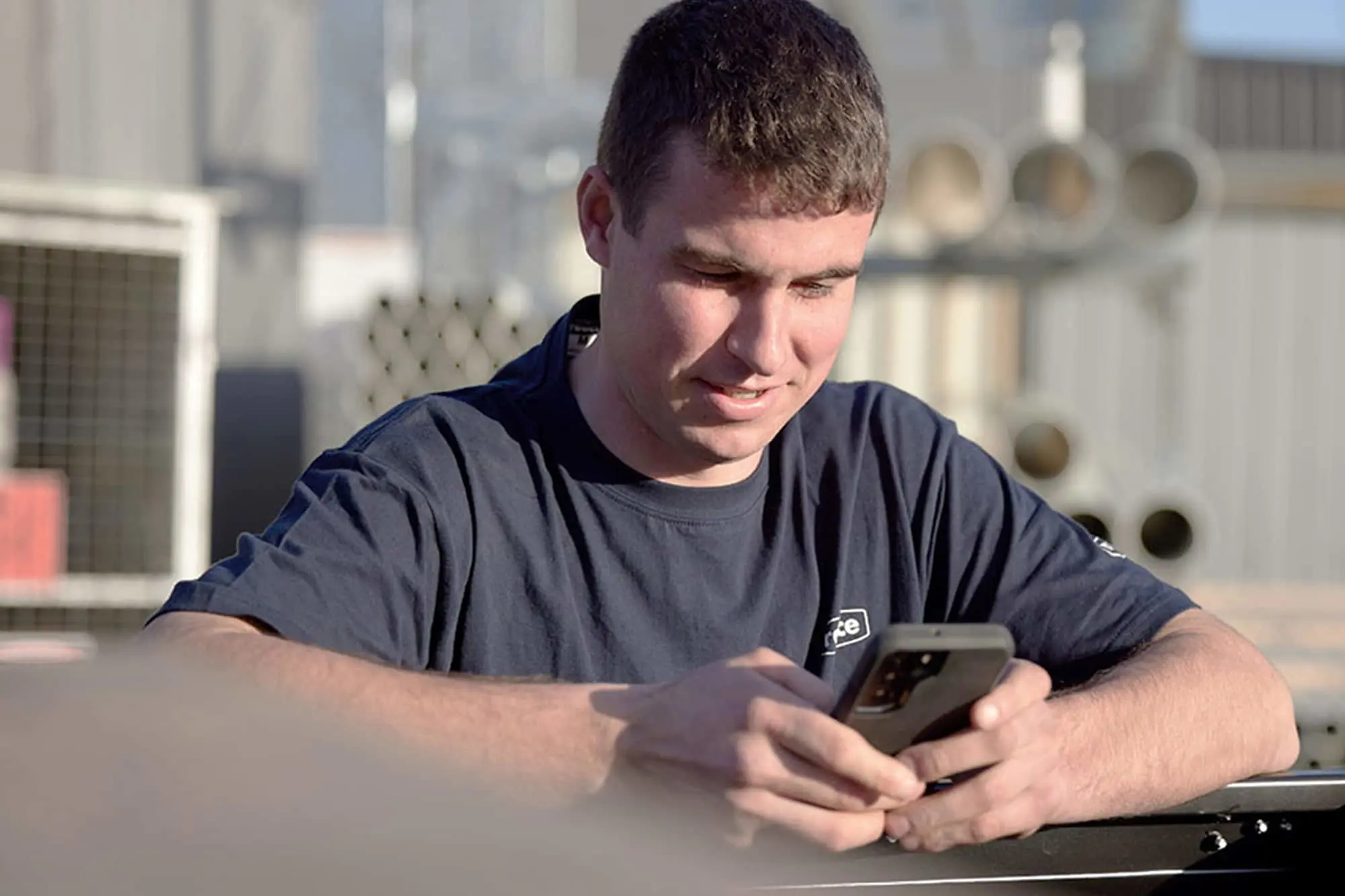 Man in navy blue shirt looking down at smartphone outdoors with blurred building background.