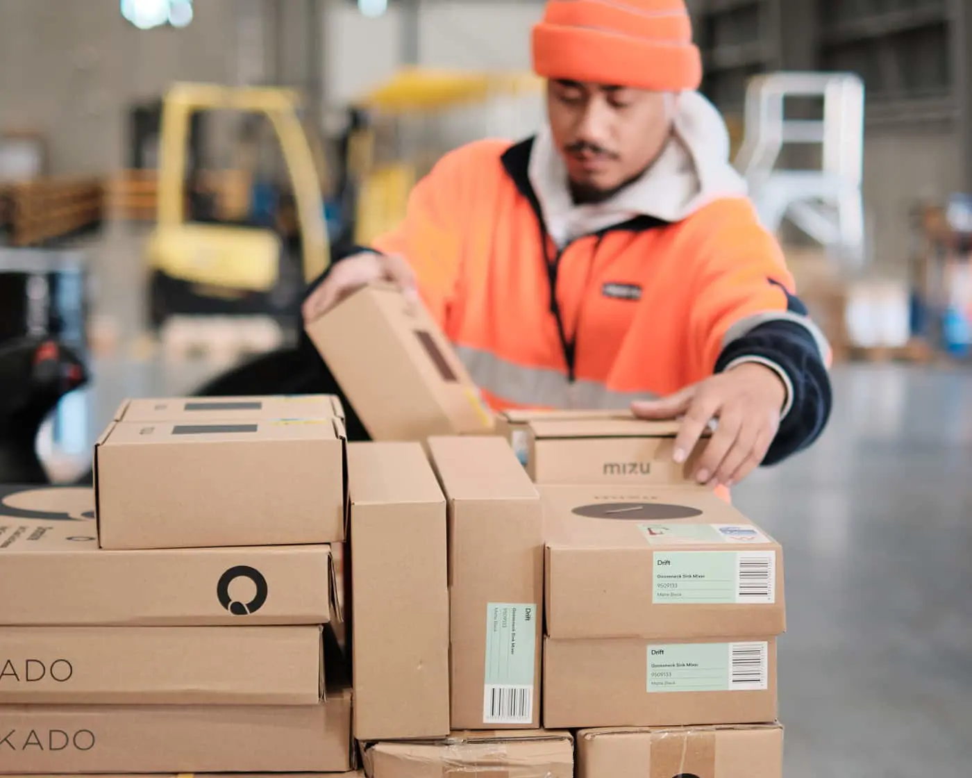 Warehouse worker in orange safety jacket and beanie sorting cardboard boxes with shipping labels in a warehouse.