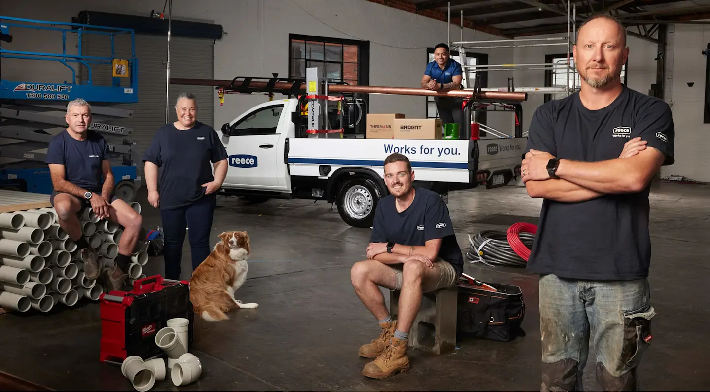 Four workers in navy shirts and a dog pose in a warehouse with a white work van, pipes, and equipment.