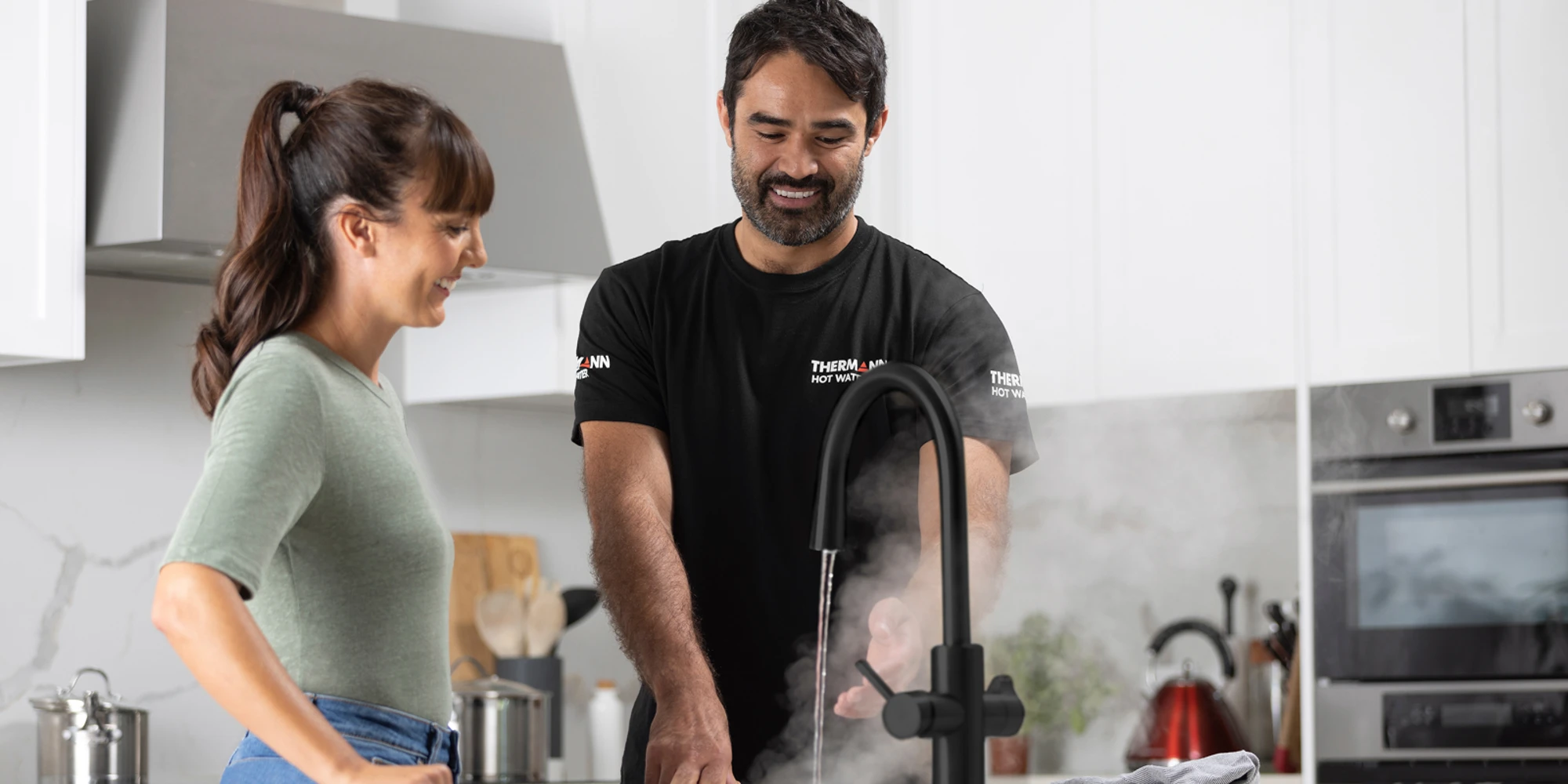 Technician in black uniform demonstrating a black kitchen faucet to a woman in a modern white kitchen.