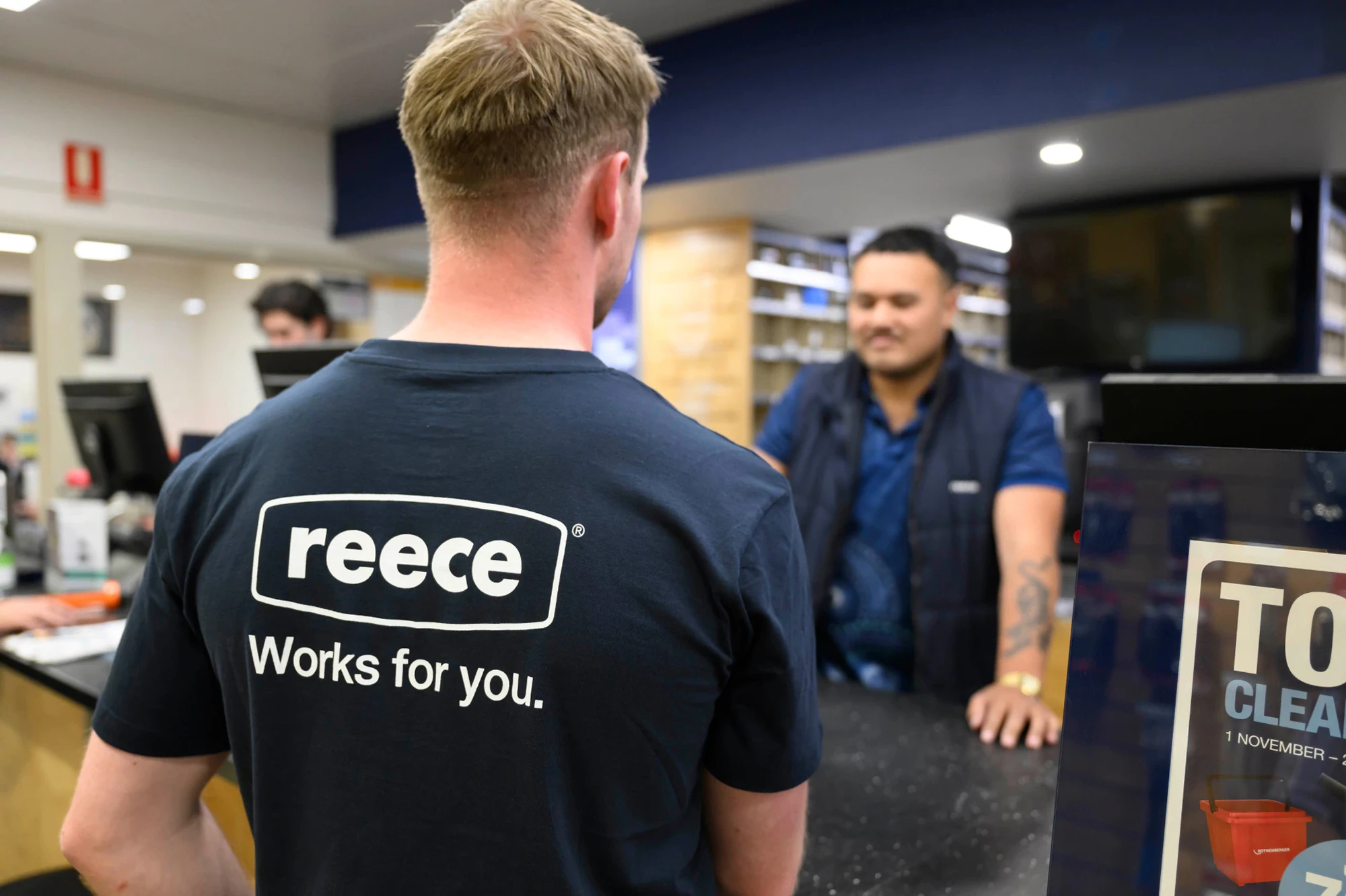 A Reece staff member in a branded black t-shirt stands at a counter speaking with a customer inside a Reece store.