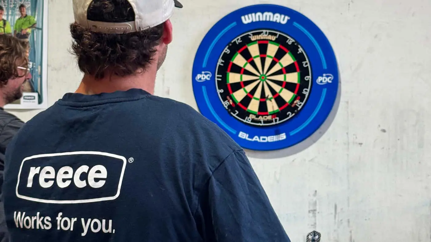 Person in dark shirt and white cap facing a Winmau dartboard with blue surround mounted on white wall.