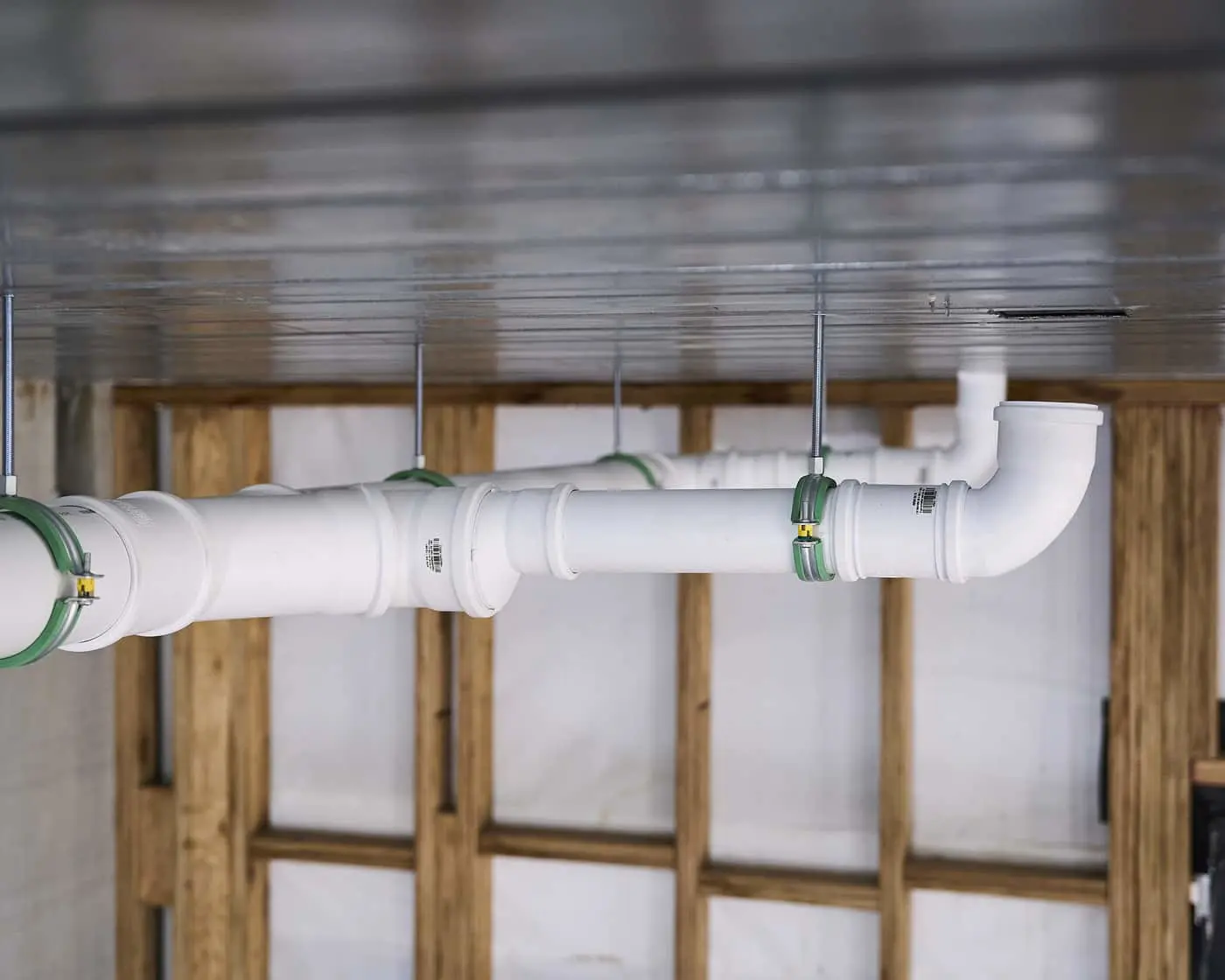 White PVC pipes with green clamps attached to wooden floor joists in an unfinished basement or crawl space.