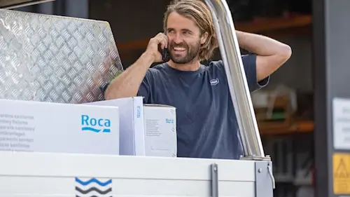 Man carrying a metal sheet and Roca branded box while talking on phone outdoors.