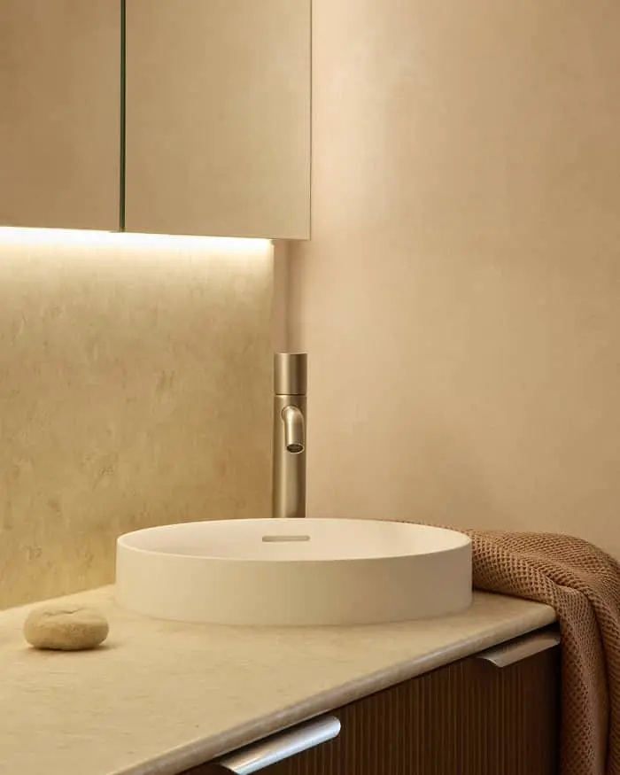 White round sink with brushed gold faucet on beige countertop beneath wall-mounted cabinet with under-cabinet lighting