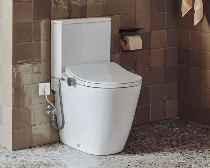 White modern toilet with bidet attachment in bathroom with brown tiled walls and speckled floor.