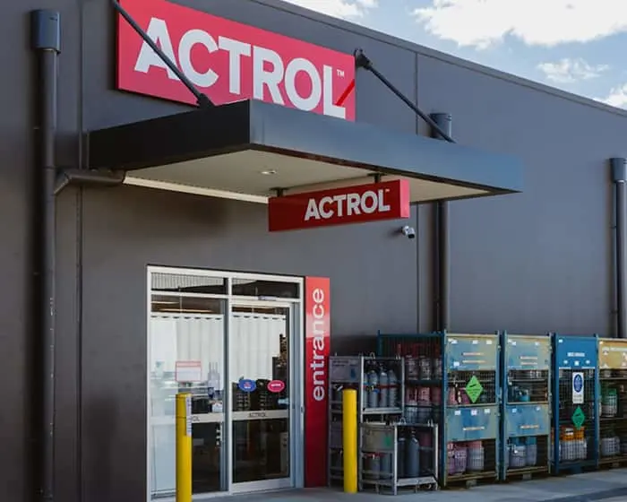 Modern gray building exterior with red ACTROL signage above entrance and gas cylinders in cages on right side.