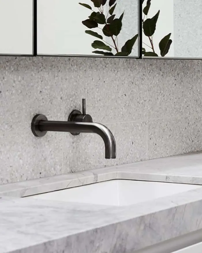 Modern bathroom with black wall-mounted faucet, white marble countertop, and framed mirror with plant reflection.