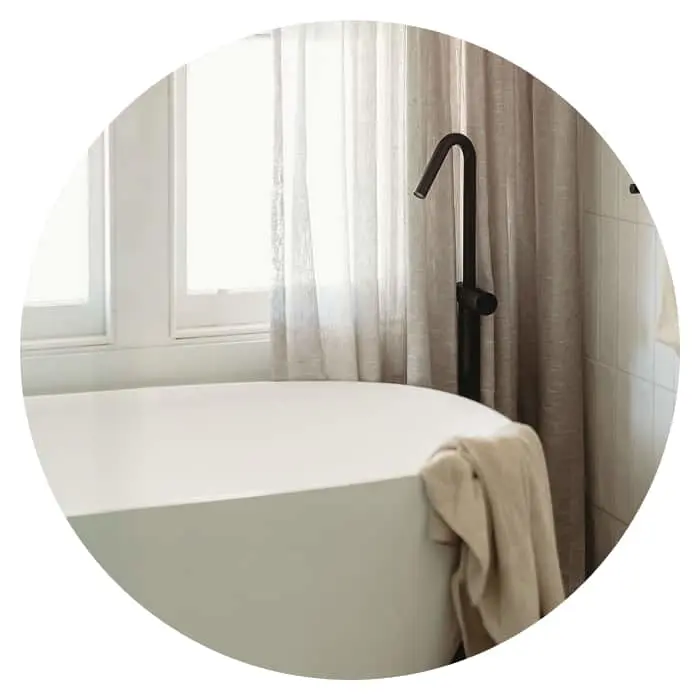 White freestanding bathtub with black faucet, beige towel draped over edge, sheer curtains and window in background.