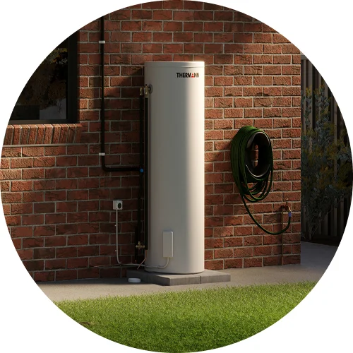 White cylindrical Thermann hot water heater installed against red brick wall with green garden hose mounted nearby