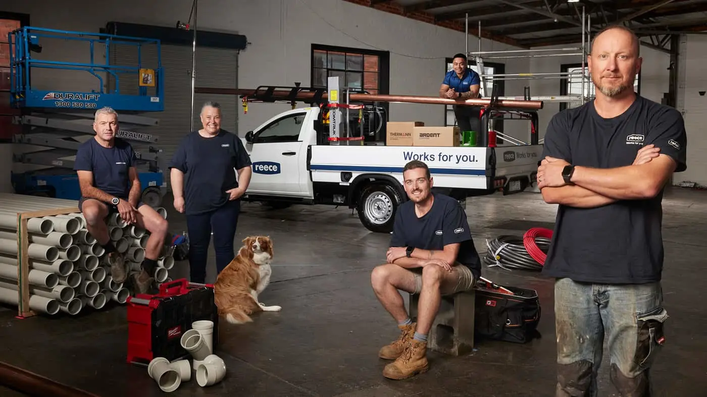 Four workers and a dog pose in a warehouse with a white work truck, blue lift, and plumbing supplies.