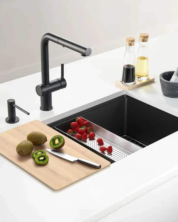 Black kitchen sink with matte black faucet, cutting board with kiwis, and colander with strawberries on white counter