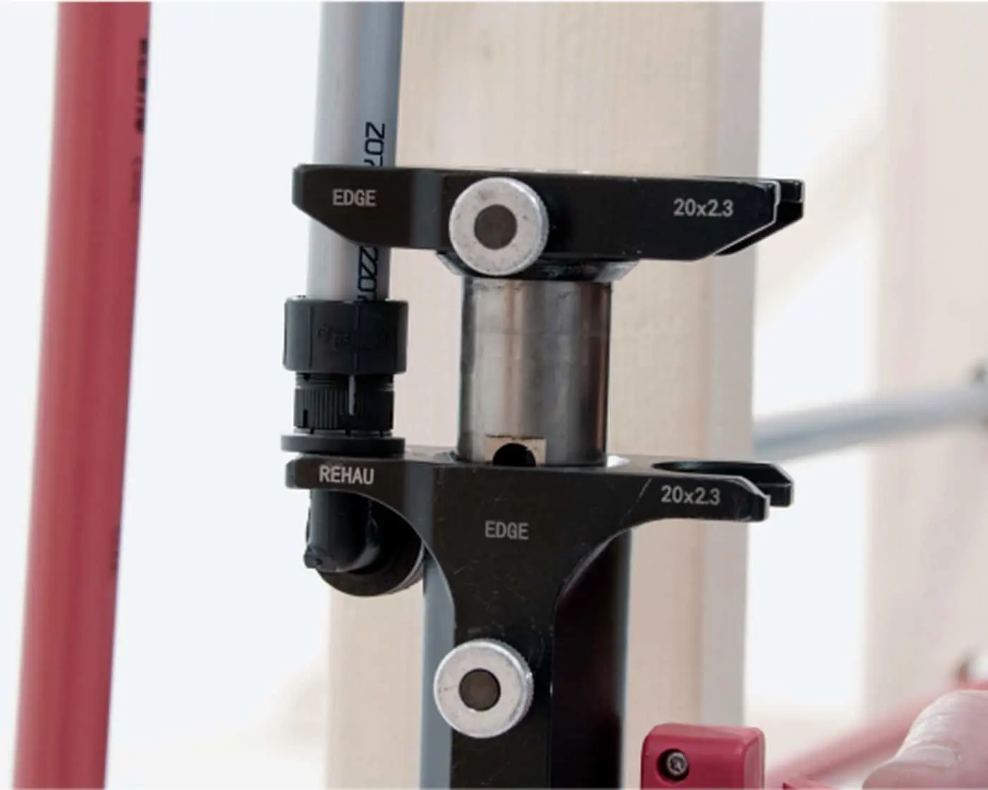 Black metal pipe clamping tool labeled REHAU with 20x2.3 markings, positioned on gray pipes against a light background.