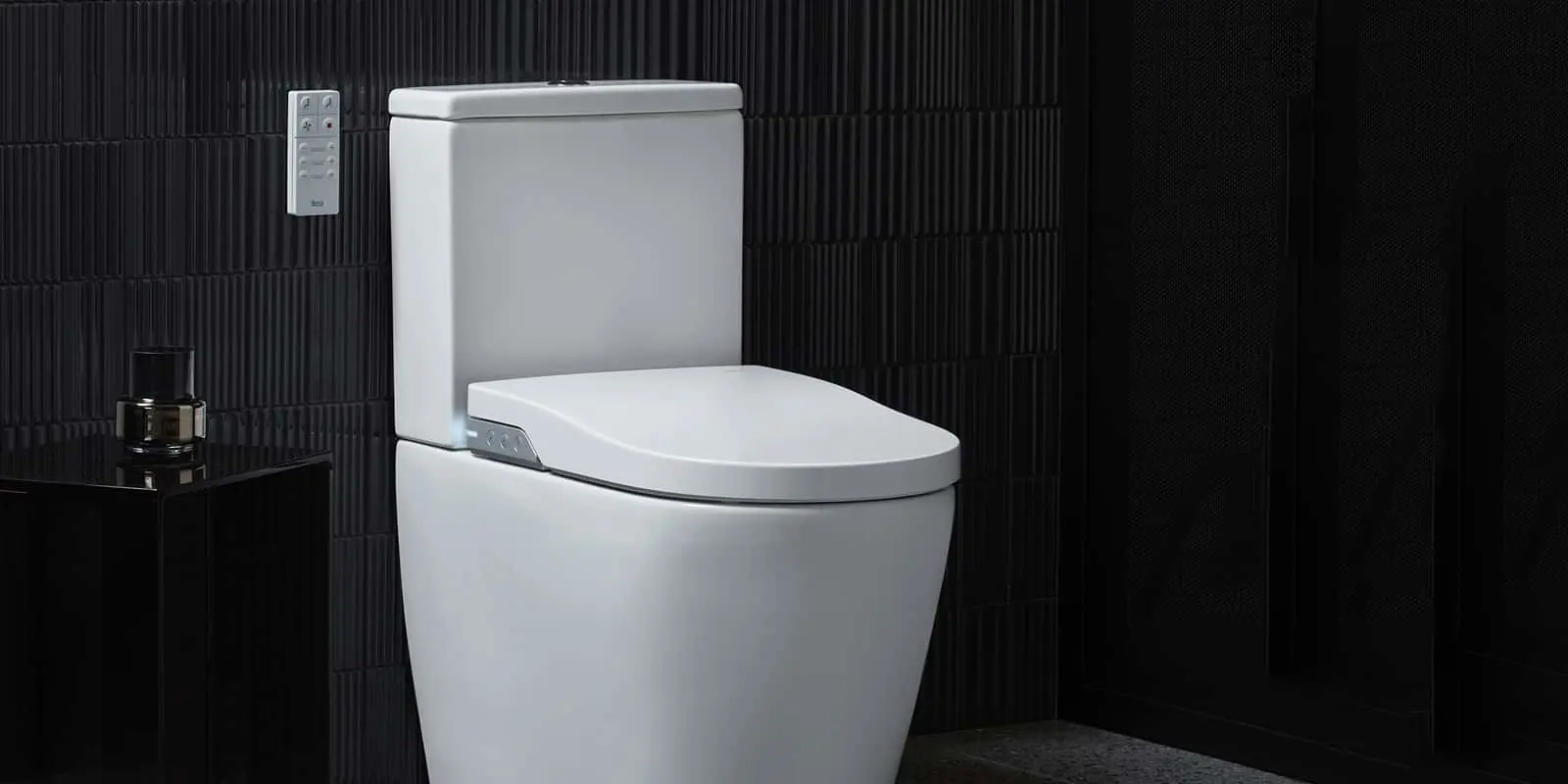 White modern toilet with electronic bidet seat and wall-mounted remote control against black textured tile wall.