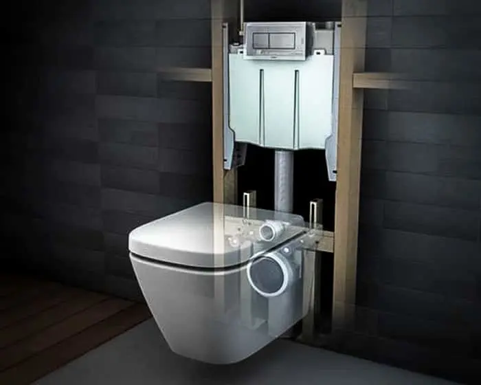 Wall-mounted toilet with exposed in-wall tank and plumbing system against dark tiled bathroom wall