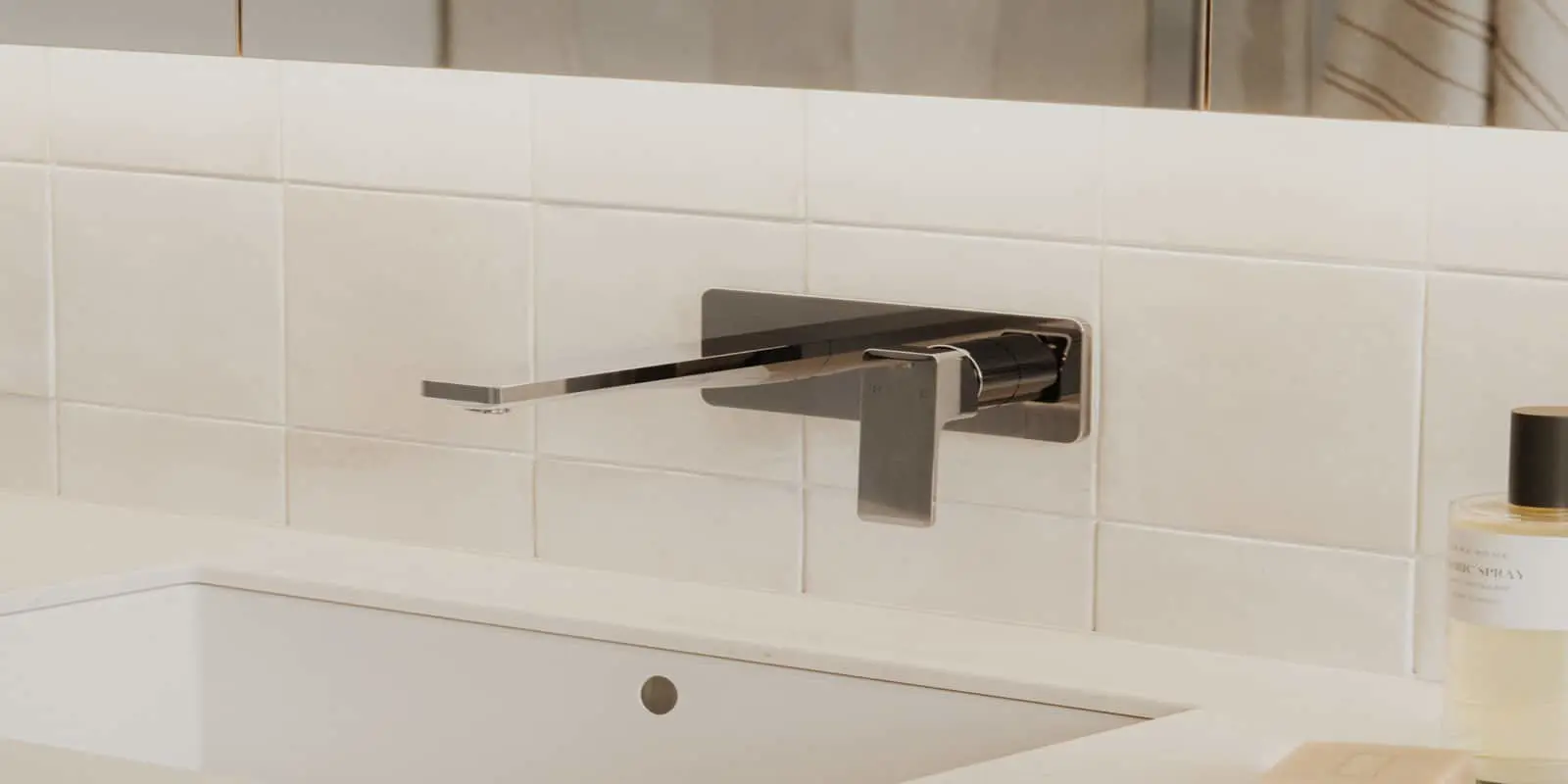 Modern wall-mounted faucet with brushed metal finish above white sink against white tile backsplash.