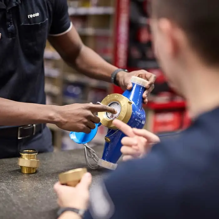 Two people examining a blue plumbing fixture with brass fittings on a workbench.