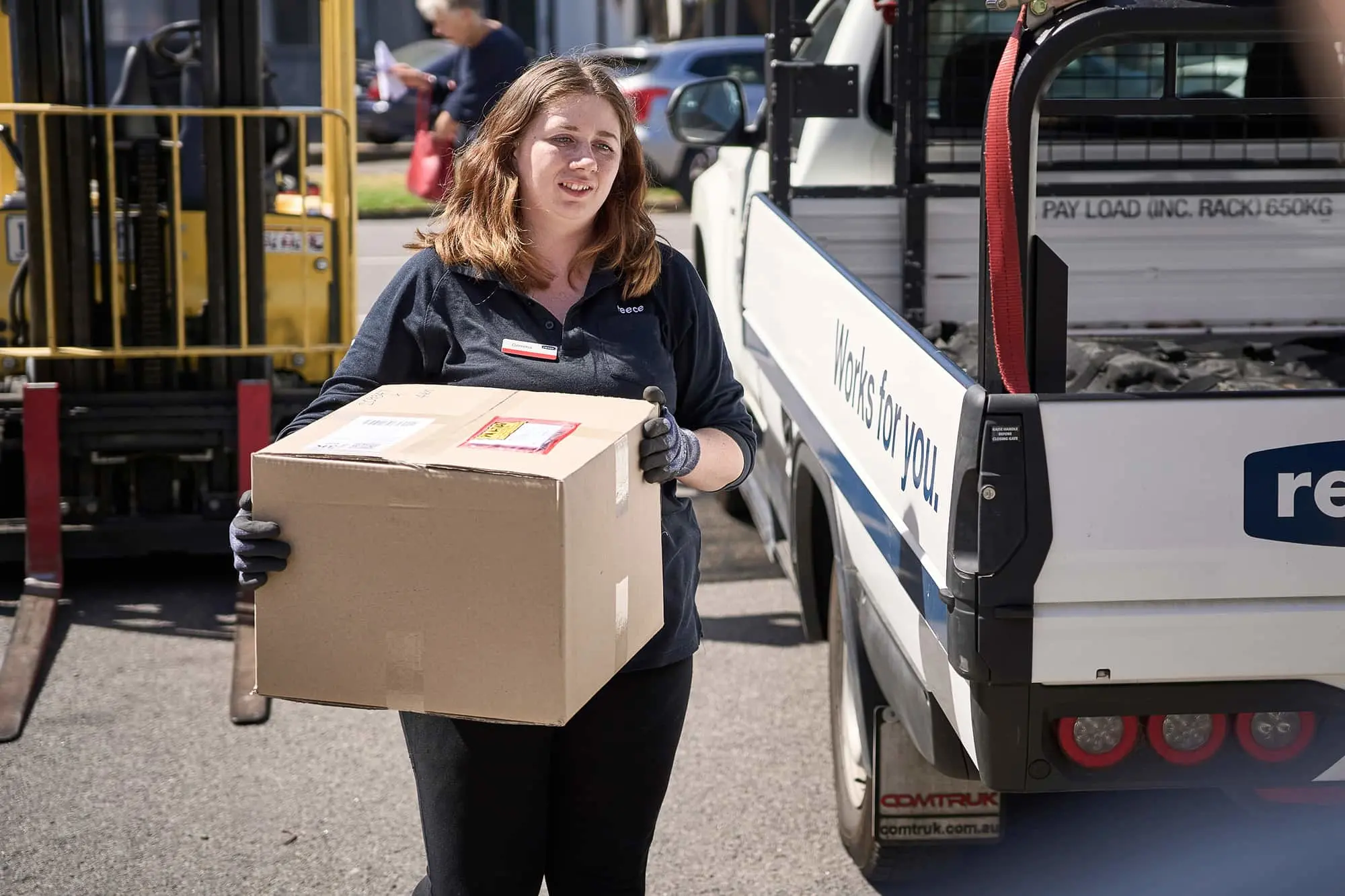 Woman in dark uniform carrying cardboard box near white delivery truck with yellow forklift in background.