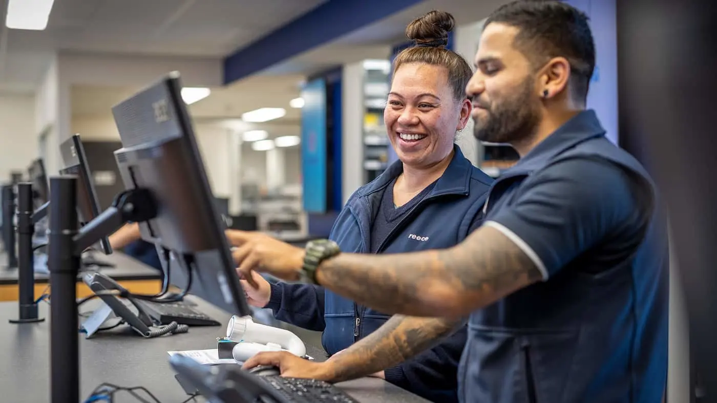 Two people in blue uniforms working together at a computer desk, woman smiling while man points at screen.