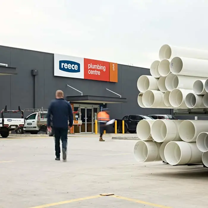 Person walking toward Reece Plumbing Centre storefront with stacked white pipes in foreground and parking lot.