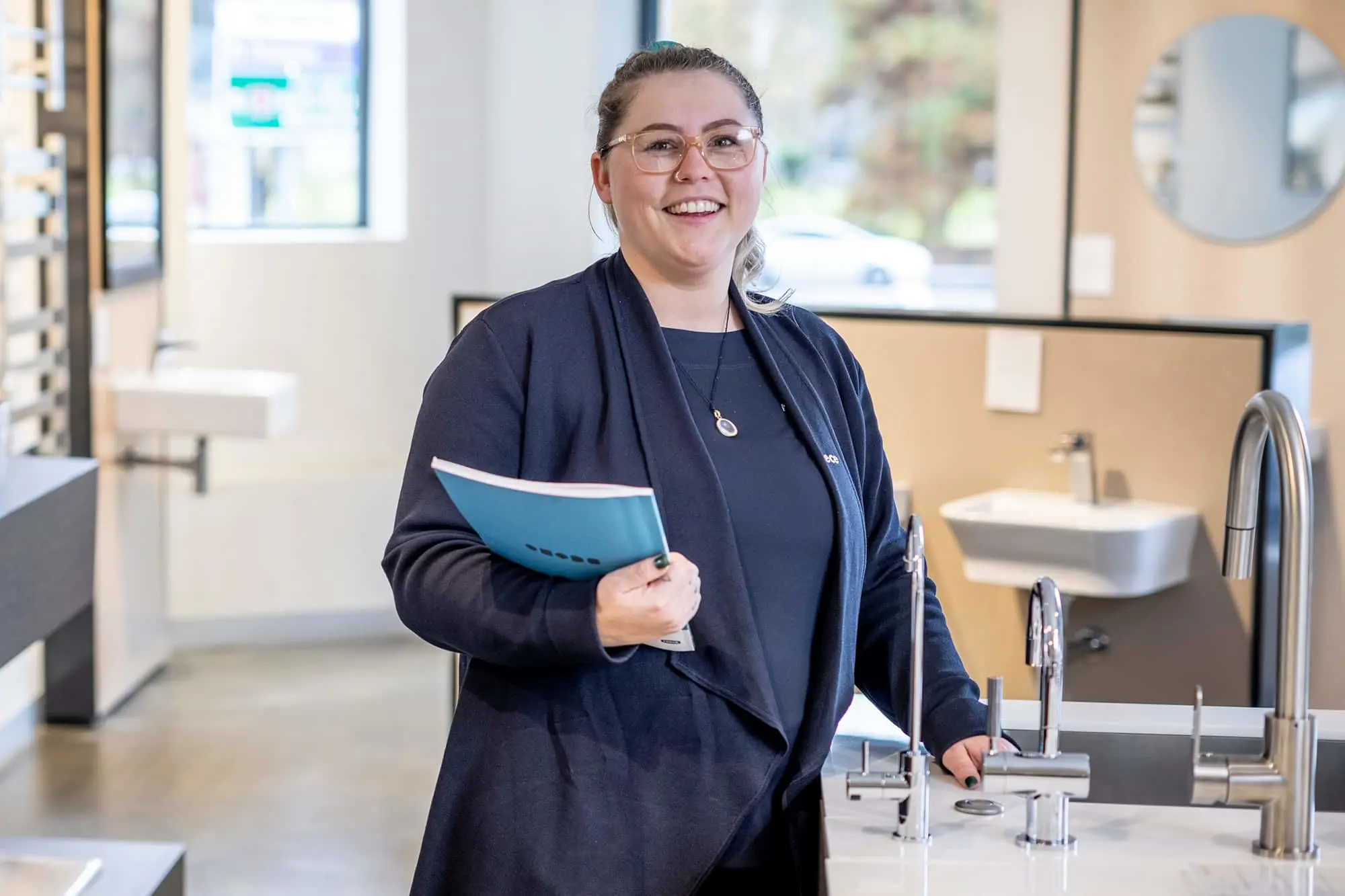 Woman in navy blazer and glasses holding blue folder in bathroom showroom with sinks and fixtures displayed.