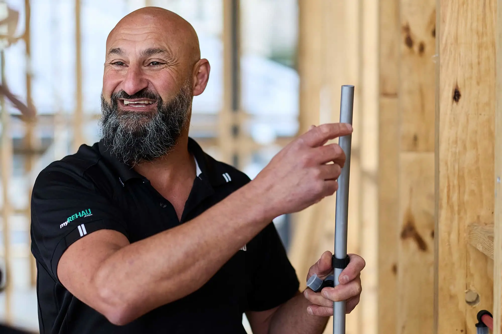 Smiling bald man with beard in black polo shirt holding a metal pole against wooden wall in workshop setting.