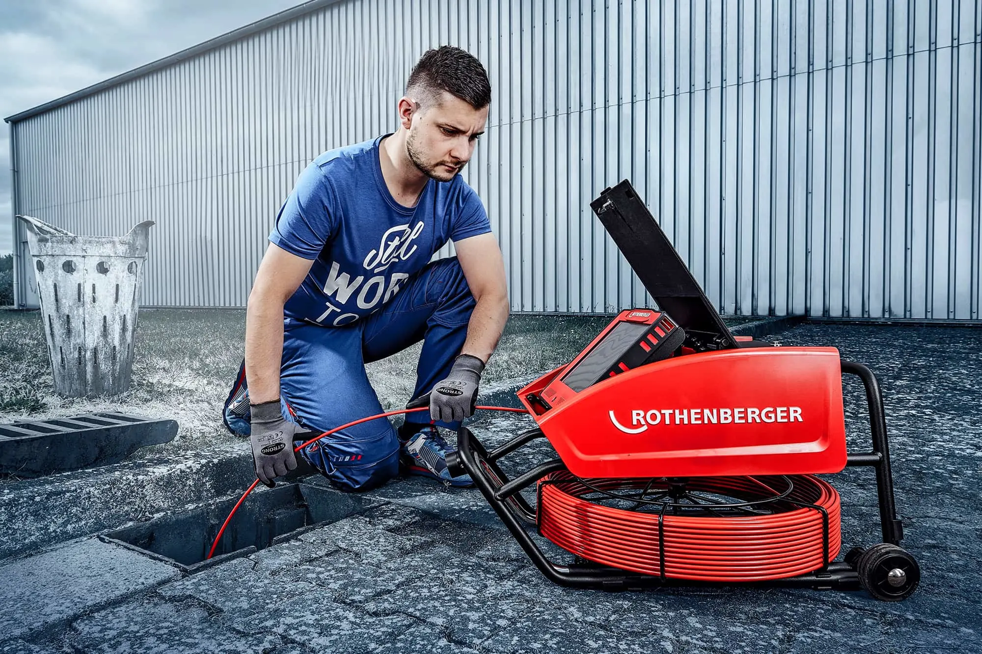 Man in blue shirt kneeling beside red Rothenberger pipe inspection equipment on pavement near metal building.