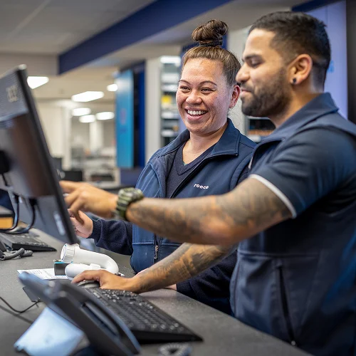 Two smiling employees in navy blue uniforms working together at a computer workstation in an office setting.