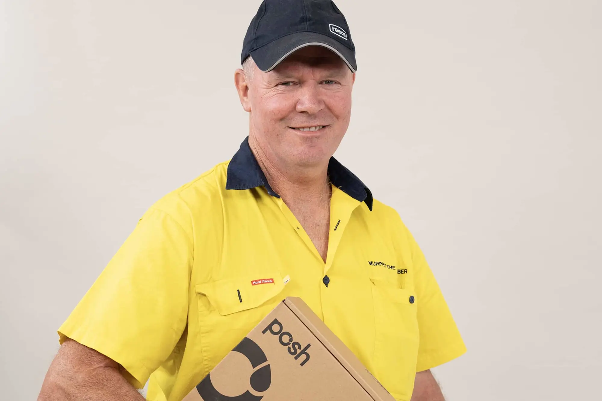 Man wearing dark cap and yellow work shirt with dark collar holding a cardboard box labeled posh.