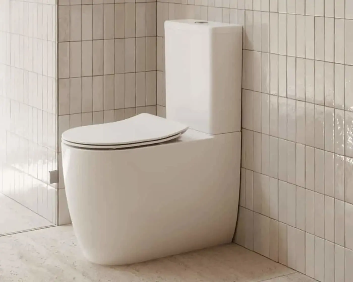 White modern toilet with closed lid against beige tiled bathroom wall next to glass shower door.